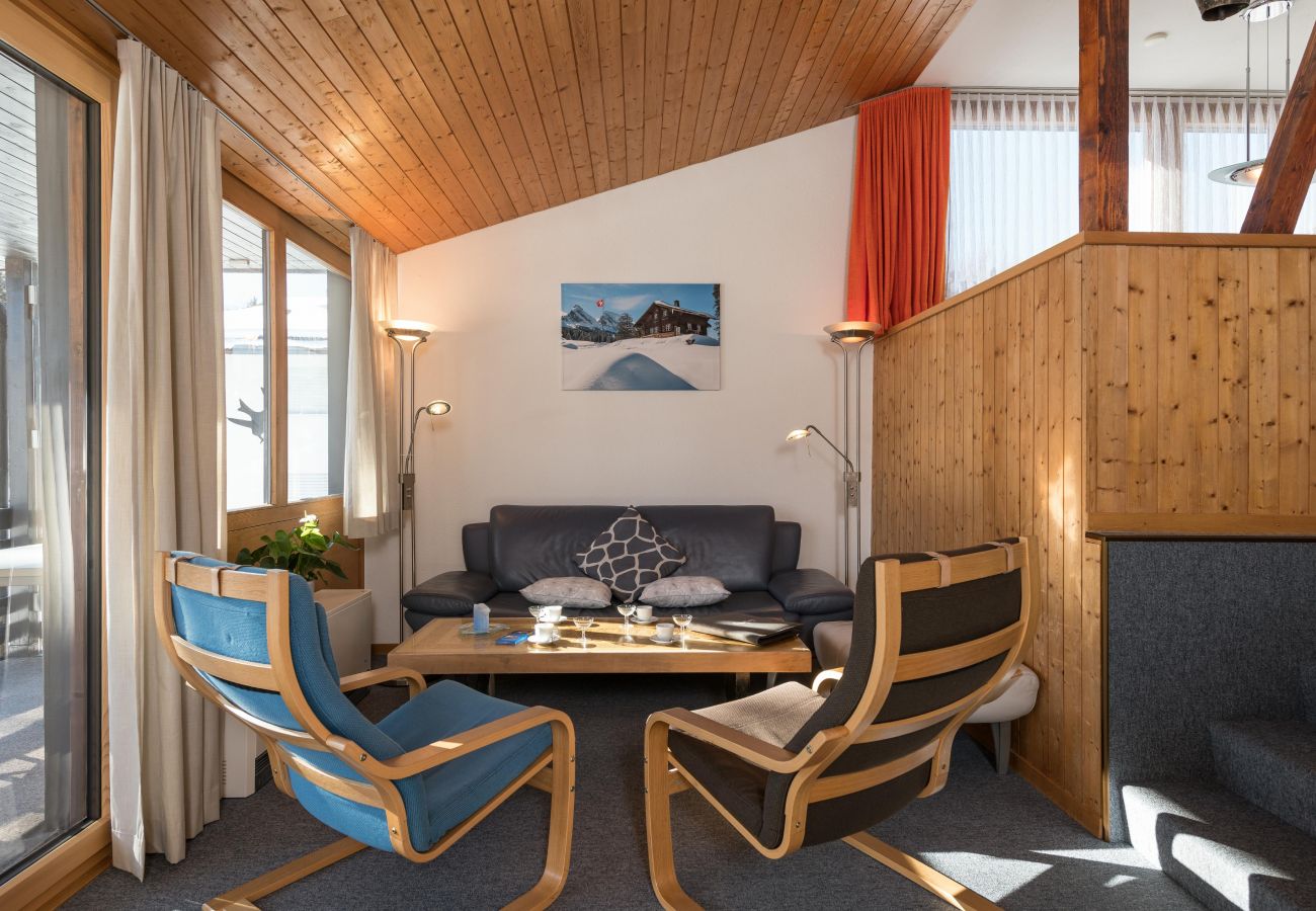 Séjour chaleureux avec plafond en bois, grandes fenêtres, canapé et deux fauteuils.