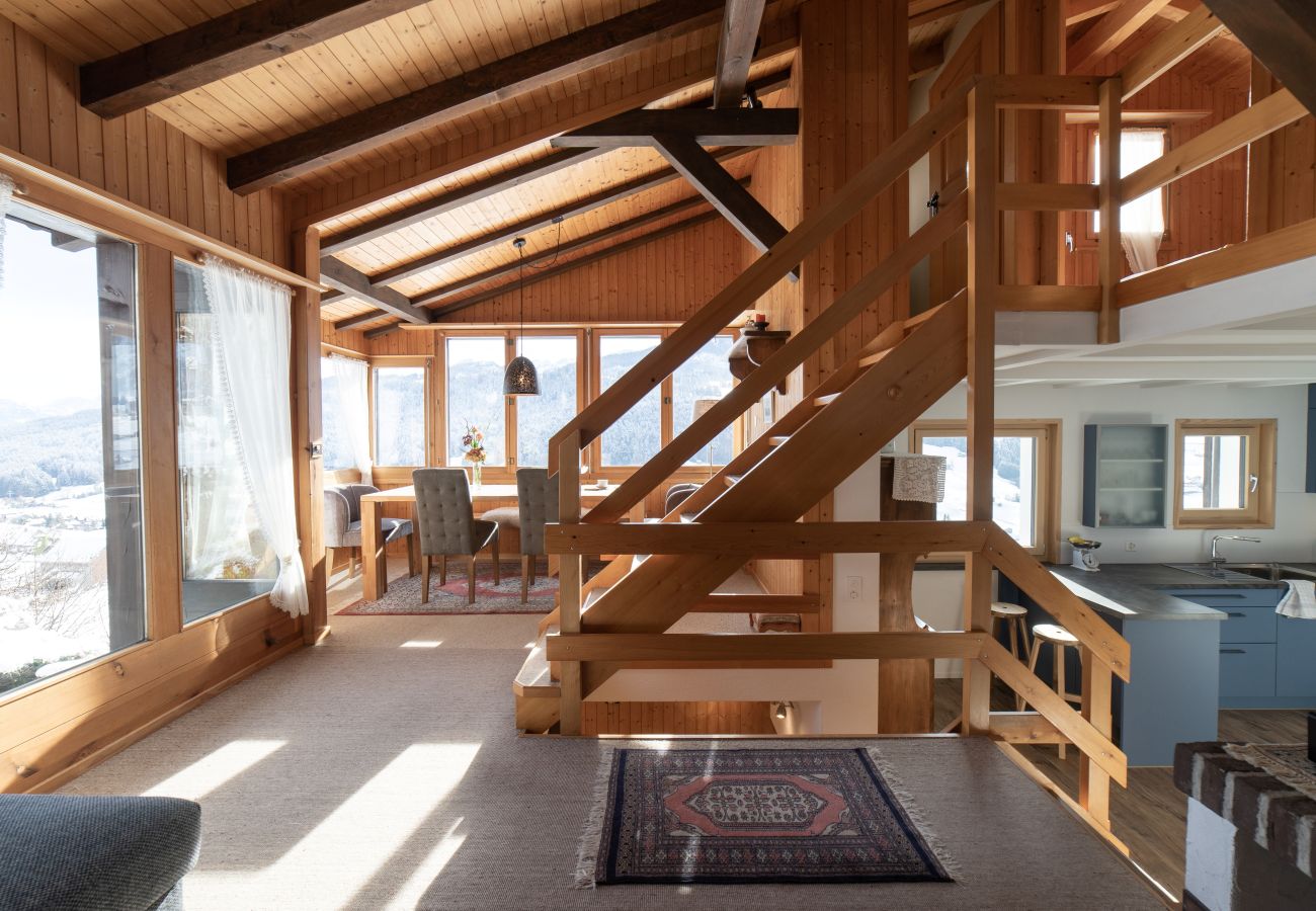 Heller Wohnbereich mit viel Holz, offener Treppe, grossen Fenstern und Blick auf die Berglandschaft.