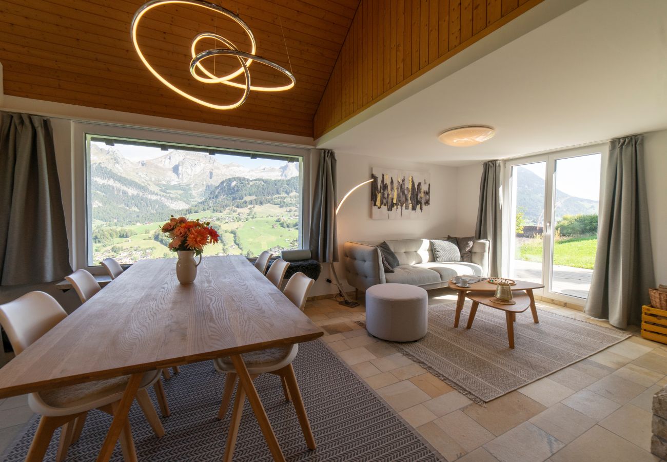 Offener Wohn- und Essbereich mit Holztisch, Sofa, grossen Fenstern und Panoramablick auf die Berge.